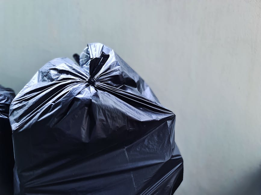 Three large black plastic garbage bags filled with waste are positioned on the pavement in front of a black metal fence, near the curbside of a street. The bags are lumpy and bulging, indicating they
