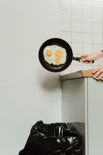 A person holding a small frying pan with a cooked breakfast consisting of a fried egg, with the yolk resembling a face, is positioned near a white tiled wall and is partially visible on the right side